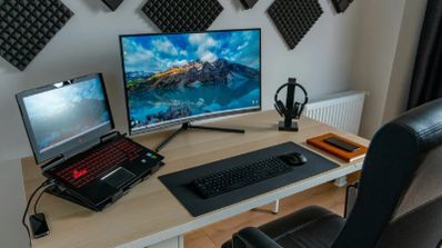 Escritorio de trabajo con un portátil gaming conectado a un monitor externo de gran tamaño, teclado completo y ratón sobre una alfombrilla negra.