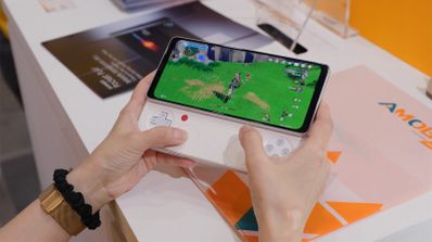 Persona sujetando una consola portátil blanca Ayaneo Pocket DMG con pantalla panorámica ejecutando un videojuego de mundo abierto.