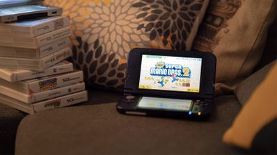 Consola portátil Nintendo 3DS abierta mostrando el menú de New Super Mario Bros. 2 junto a una pila de juegos físicos sobre un sofá.