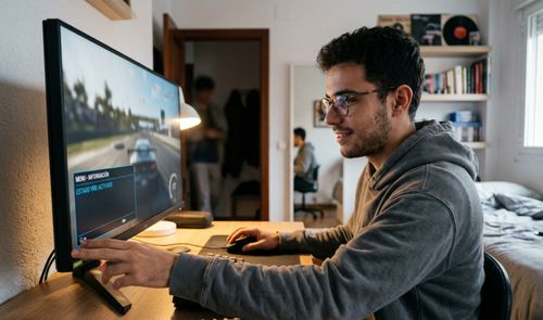 Joven con gafas configurando un monitor gaming de pantalla ancha mientras juega a un simulador de conducción en su escritorio.