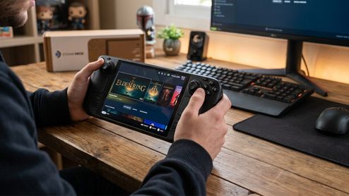 Primer plano de una persona sosteniendo la consola portátil Steam Deck frente a un escritorio de PC con la biblioteca de juegos abierta.