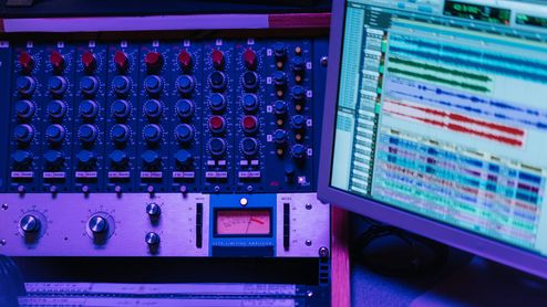 Mesa de mezclas analógica con diales rojos y azules junto a un monitor de ordenador que muestra un software de edición de audio profesional.