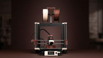 Impresora 3D con dos bobinas de filamento en la parte superior en colores beige y marrón sobre un fondo oscuro.