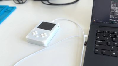 Consola portátil pequeña de color blanco translúcido conectada mediante un cable USB a un ordenador portátil sobre una mesa de trabajo.