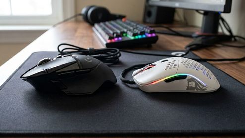 Comparativa entre un ratón gaming ergonómico negro de perfil robusto y un ratón ultraligero blanco con diseño de panal sobre una alfombrilla.