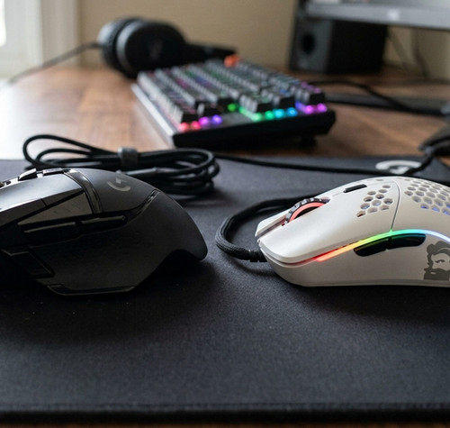 Comparativa entre un ratón gaming ergonómico negro de perfil robusto y un ratón ultraligero blanco con diseño de panal sobre una alfombrilla.