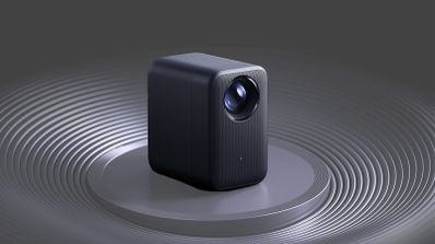 Proyector compacto de color gris oscuro con lente frontal circular sobre una base con relieve de ondas concéntricas.
