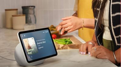 Altavoz inteligente Echo Show en una encimera de cocina reproduciendo música mientras una persona cocina al fondo.