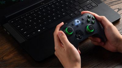 Manos sosteniendo un mando inalámbrico 8BitDo con detalles en verde frente a un ordenador portátil