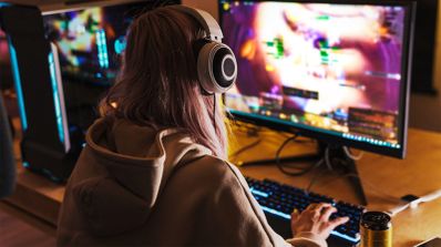 Vista posterior de una mujer con auriculares frente a un monitor gaming con teclado retroiluminado.
