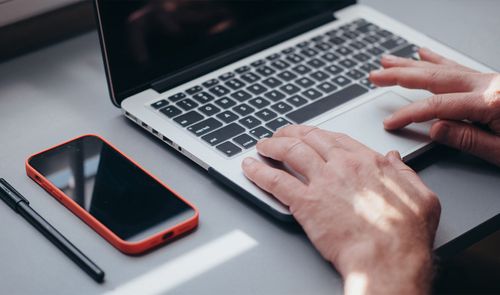 Manos de una persona utilizando el panel táctil de un ordenador portátil junto a un teléfono móvil con funda roja sobre una mesa de trabajo.