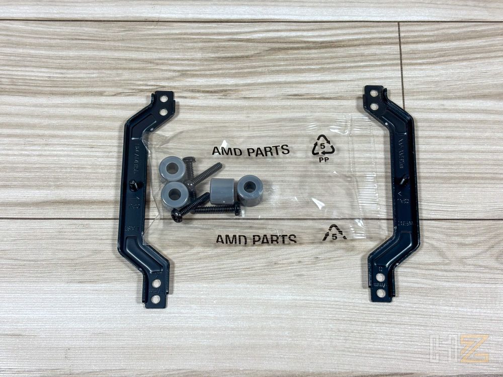 Soportes de montaje metálicos negros junto a una bolsa transparente con tornillos y espaciadores etiquetada como AMD PARTS.
