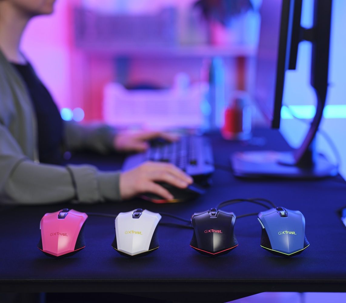 Cuatro ratones gaming Trust GXT 109W en colores rosa, blanco, negro y azul sobre una alfombrilla oscura frente a un setup de ordenador.
