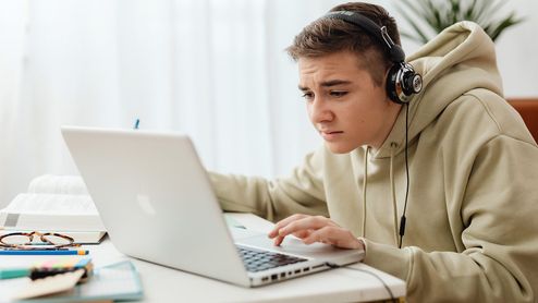 Joven con auriculares concentrado frente a su ordenador portátil en un escritorio con libros.