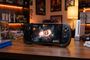 Consola portátil ARC-X1 ejecutando un juego de acción sobre un escritorio de madera rodeado de juegos y consolas clásicas.