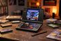 Consola portátil Yoniq Duo con doble pantalla sobre una mesa rodeada de cartuchos de videojuegos clásicos y revistas.