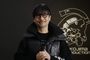 Retrato de Hideo Kojima con gafas y chaqueta oscura frente al logotipo de Kojima Productions.