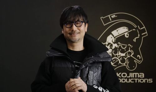 Retrato de Hideo Kojima con gafas y chaqueta oscura frente al logotipo de Kojima Productions.