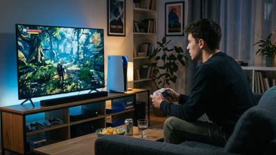 Usuario sentado en un sofá manejando un mando frente a un televisor que muestra un videojuego y una consola PS5 al lado.