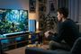 Usuario sentado en un sofá manejando un mando frente a un televisor que muestra un videojuego y una consola PS5 al lado.