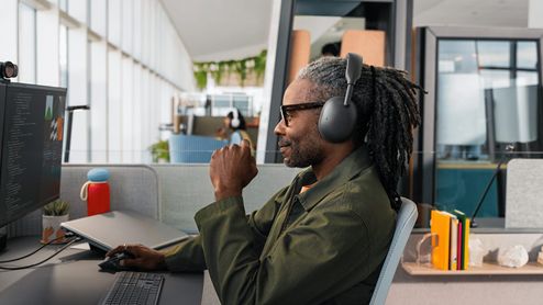 Hombre con rastas trabajando en una oficina luminosa con los auriculares Logitech Zone Vibe Pro puestos mientras mira el monitor.