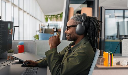 Hombre con rastas trabajando en una oficina luminosa con los auriculares Logitech Zone Vibe Pro puestos mientras mira el monitor.