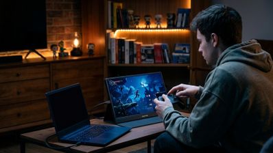 Persona jugando con un mando frente a un monitor gaming portátil conectado a un ordenador en una habitación con iluminación cálida.