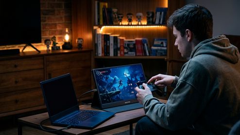 Persona jugando con un mando frente a un monitor gaming portátil conectado a un ordenador en una habitación con iluminación cálida.