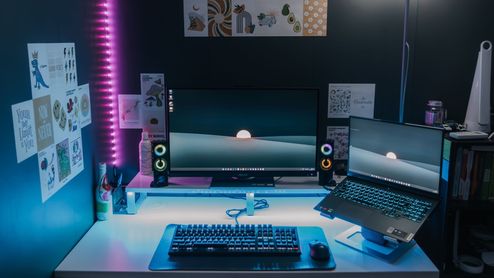 Configuración de PC gaming con monitor, ordenador portátil sobre soporte y periféricos retroiluminados en un entorno con luces LED.