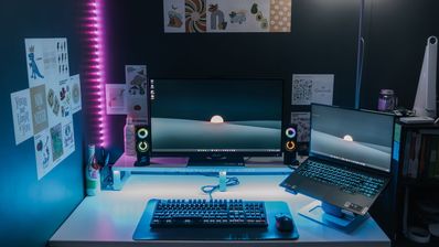 Configuración de PC gaming con monitor, ordenador portátil sobre soporte y periféricos retroiluminados en un entorno con luces LED.