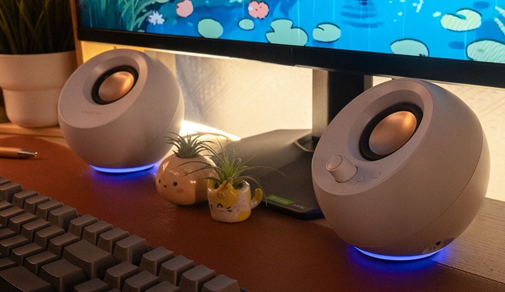 Altavoces Pebble Pro de color blanco con iluminación LED azul en la base, situados en un escritorio bajo un monitor.