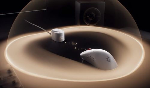 Ratón inalámbrico SteelSeries de color blanco junto a su receptor en una superficie iluminada por una cúpula de luz.