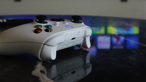 Mando inalámbrico de Xbox de color blanco sobre una mesa con una interfaz de juegos de Smart TV desenfocada al fondo.