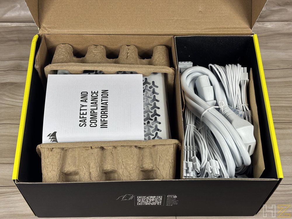 Interior de la caja de la fuente Corsair RM1200e con la unidad principal protegida por cartón reciclado y los cables blancos organizados en un compartimento lateral.