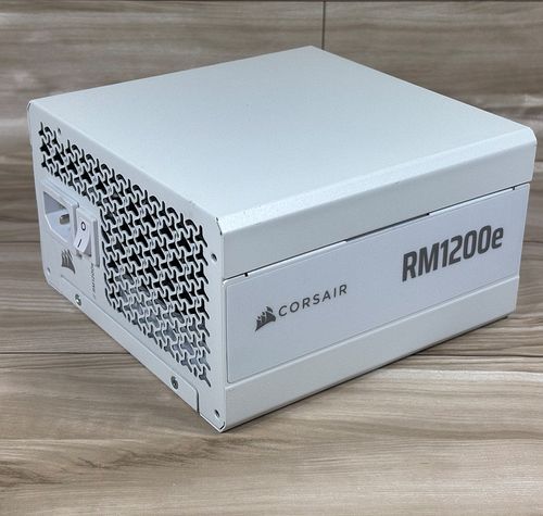 Fuente de alimentación Corsair RM1200e blanca vista en perspectiva sobre una superficie de madera.