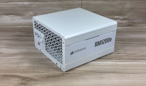 Fuente de alimentación Corsair RM1200e blanca vista en perspectiva sobre una superficie de madera.