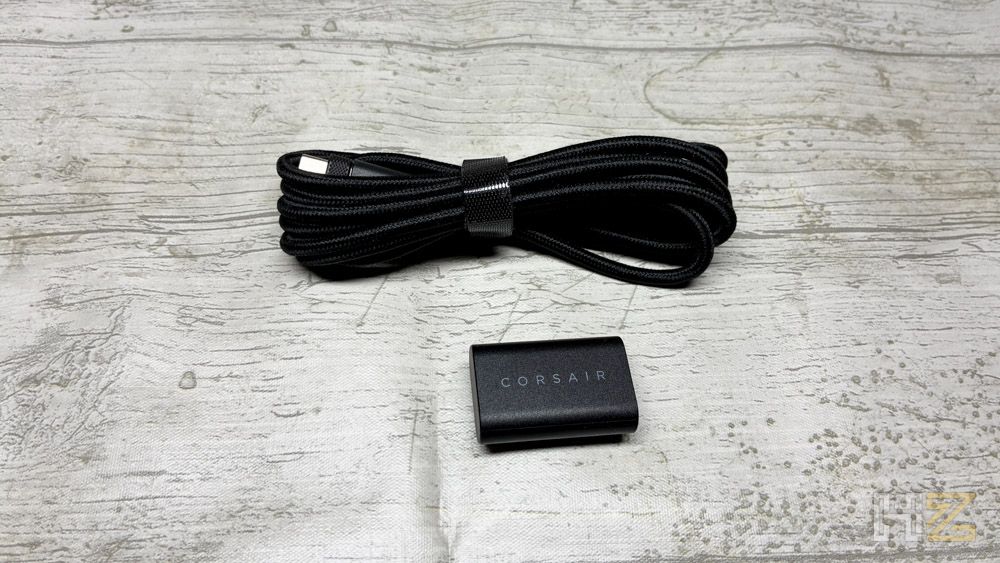 Cable de carga USB trenzado negro y adaptador inalámbrico con el logotipo de Corsair sobre una mesa de madera clara.