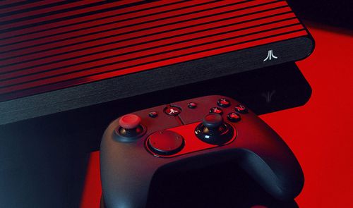 Consola Atari VCS con carcasa de rejilla y mando inalámbrico sobre una superficie oscura bajo iluminación roja.