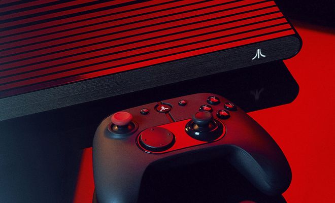 Consola Atari VCS con carcasa de rejilla y mando inalámbrico sobre una superficie oscura bajo iluminación roja.