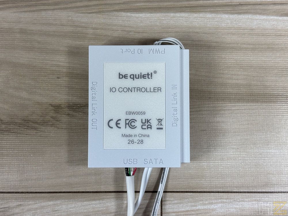 Primer plano del controlador IO blanco de be quiet! sobre una superficie de madera, mostrando sus conexiones y certificaciones.