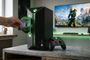 Inserción de un disco físico en una consola con diseño vertical Xbox Helix junto a un mando y una pantalla con Halo Infinite.