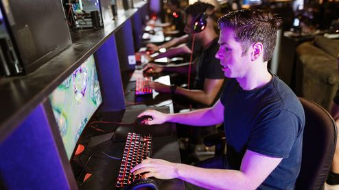 Personas sentadas frente a ordenadores en una sala de juegos LAN con teclados retroiluminados y auriculares.