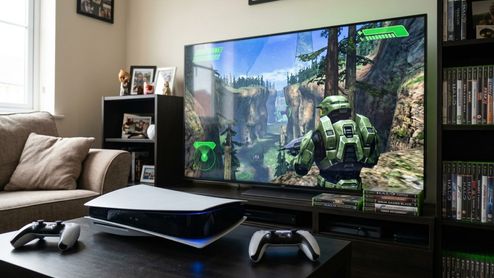 Consola PS5 con mandos DualSense frente a un televisor que muestra una partida del videojuego Halo.