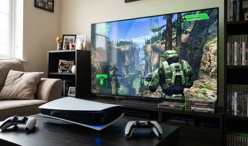 Consola PS5 con mandos DualSense frente a un televisor que muestra una partida del videojuego Halo.