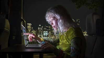 Persona señalando la pantalla táctil de un MacBook Pro en un entorno nocturno con vistas a una ciudad iluminada.