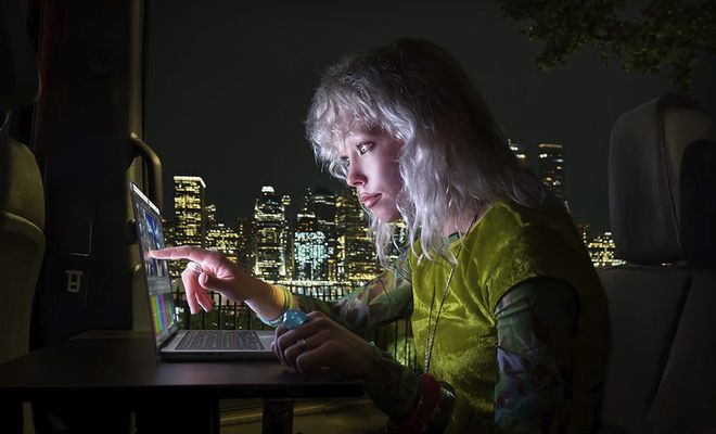 Persona señalando la pantalla táctil de un MacBook Pro en un entorno nocturno con vistas a una ciudad iluminada.