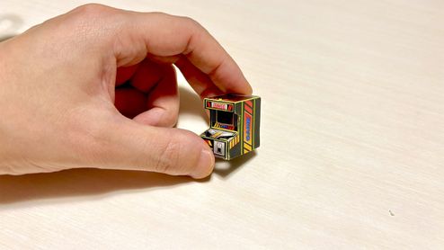 Una mano humana sujetando una réplica minúscula de una máquina recreativa arcade entre los dedos índice y pulgar.