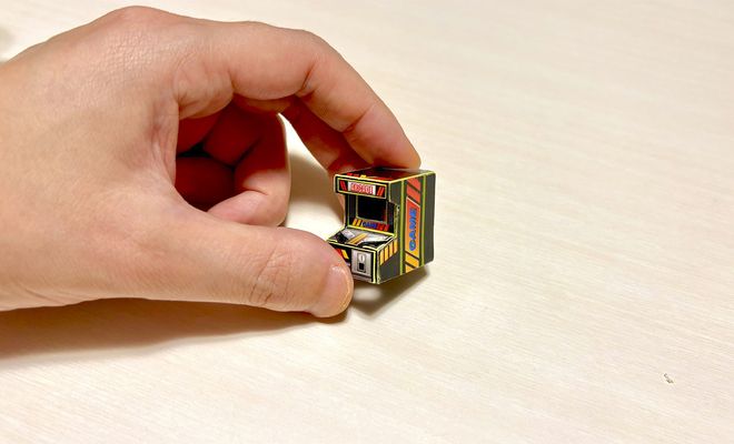 Una mano humana sujetando una réplica minúscula de una máquina recreativa arcade entre los dedos índice y pulgar.
