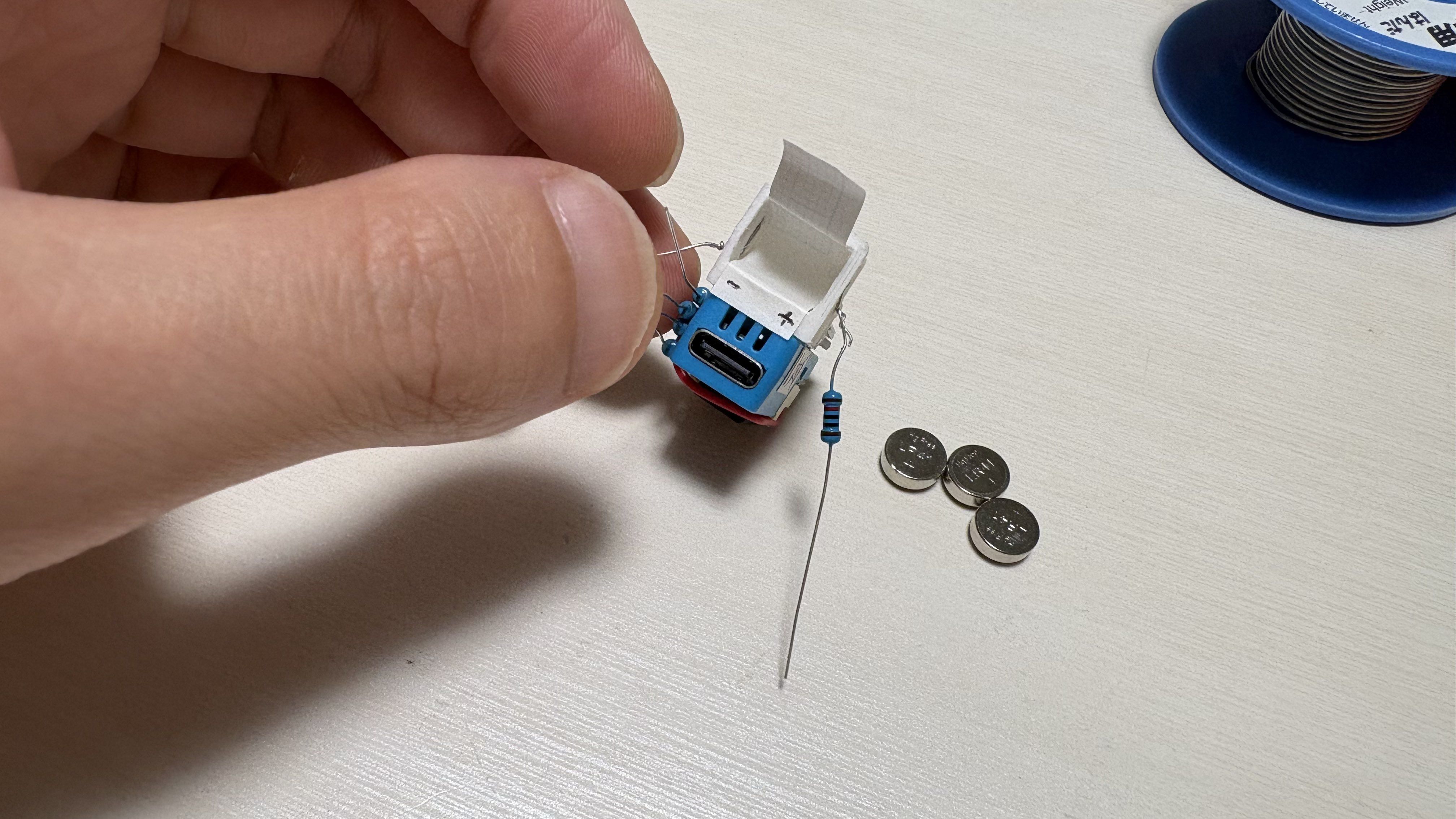 Primer plano de una mano sujetando el chasis de una mini-recreativa junto a componentes electrónicos como pilas de botón y una resistencia.