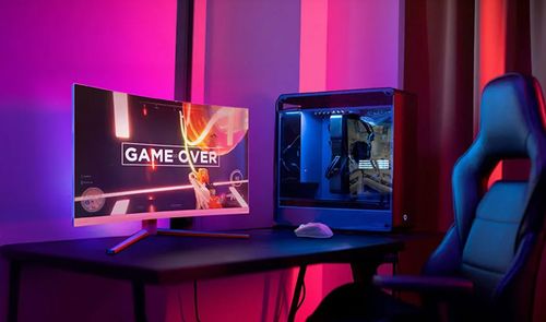 Monitor curvo Philips para videojuegos mostrando pantalla de Game Over junto a una torre de ordenador con iluminación LED azul en una habitación con luz ambiental púrpura.